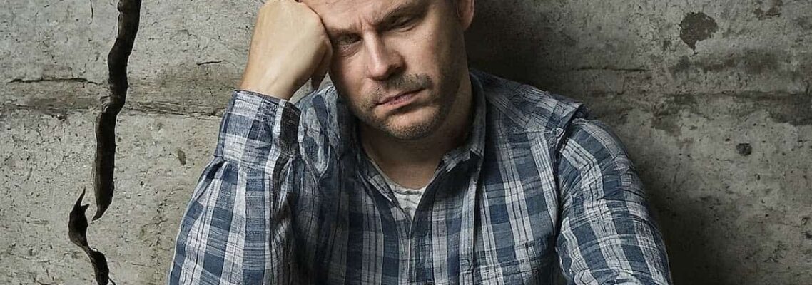 Image of a man sitting next to a large wall crack, looking regretful.