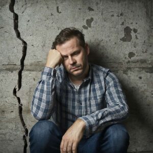 Image of a man sitting next to a large wall crack, looking regretful.